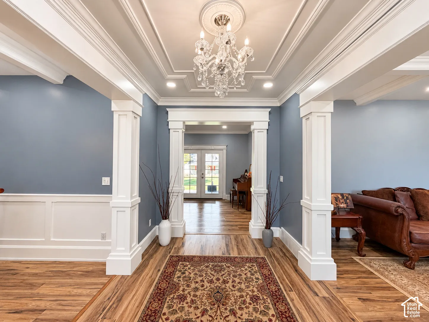 Elegant foyer showcasing coffered ceiling detail with crystal chandelier, white wainscoting and pilasters flanking the entry, blue-gray upper walls, and rich wood-finish flooring throughout