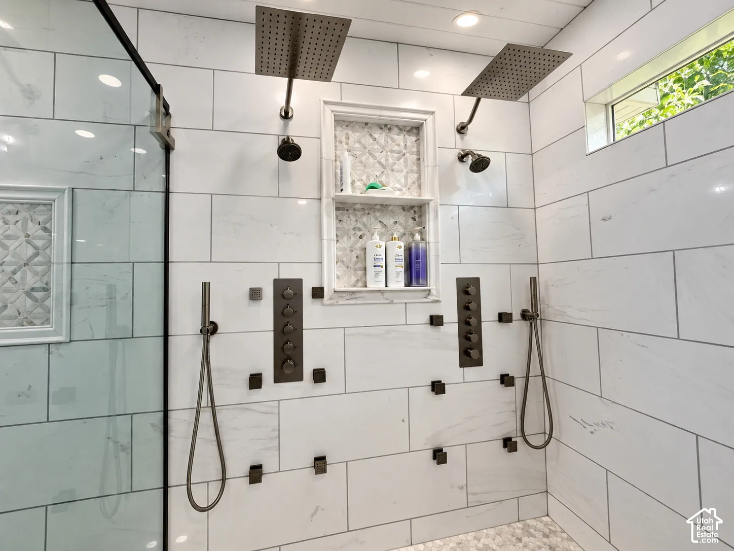 Spacious shower with large format white tile, dual rainfall showerheads, multiple body sprays, and a mosaic tile niche