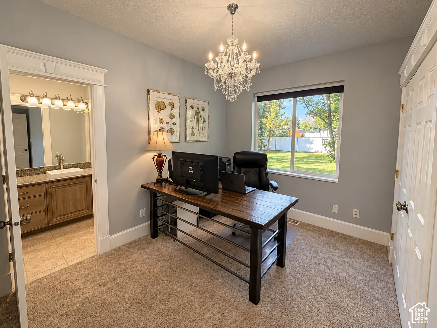 Elegant home office featuring a spacious desk with metal frame and shelving, crystal chandelier, soft gray walls, carpet flooring, and large windows with natural light