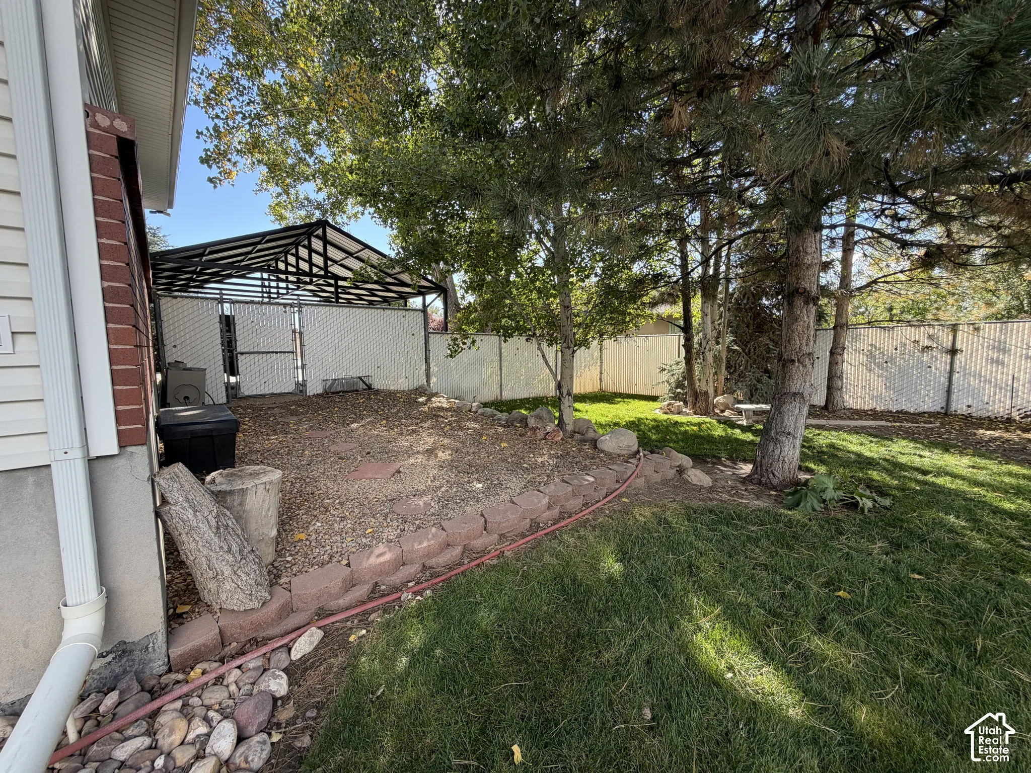 Spacious backyard oasis featuring mature shade trees, a brick-bordered planting bed, manicured lawn, metal pergola structure, and secure fencing