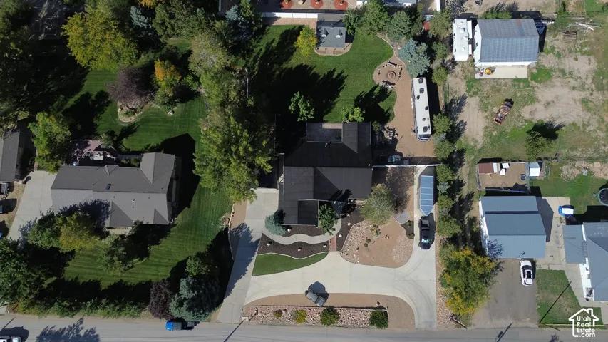 Aerial perspective featuring a paved driveway, expansive lawn, mature tree canopy, fire pit area, and rock-lined landscaping