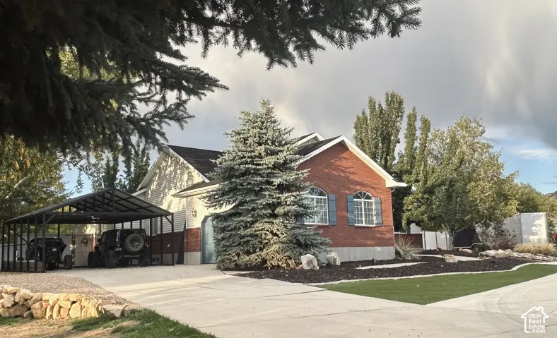 Red brick rambler with blue wooden shutters, a large covered carport for toys or extravehicles, an extended driveway and low-maintenance landscaping framed by mature trees.