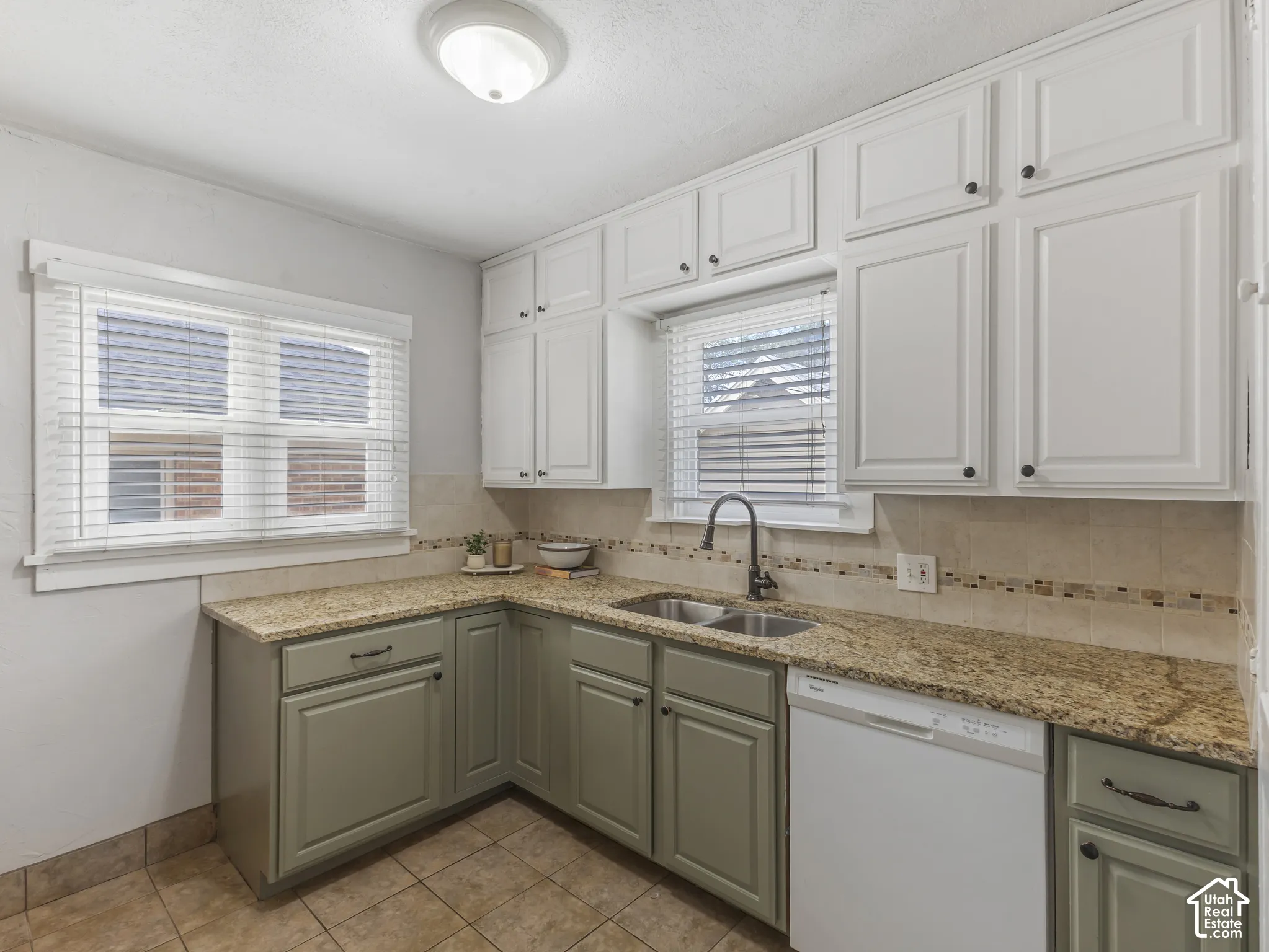 Two-tone cabinetry with upper white shaker-style cabinets and lower sage-toned cabinets, granite-finish countertops, dual basin sink with gooseneck faucet, tile backsplash, and tile flooring