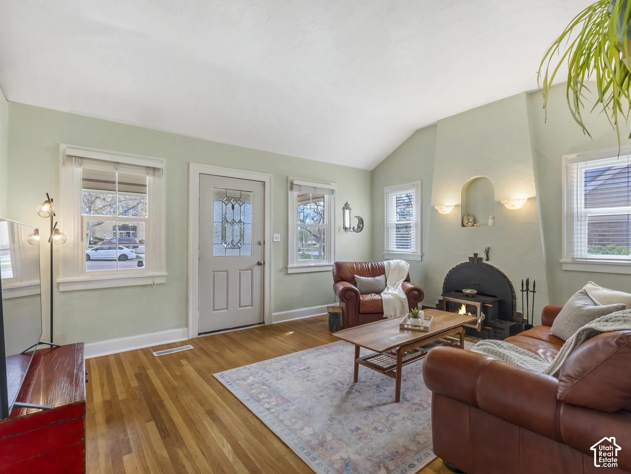 Hardwood flooring, vaulted ceiling, decorative fireplace with sconce lighting, leaded glass front door, and multiple windows