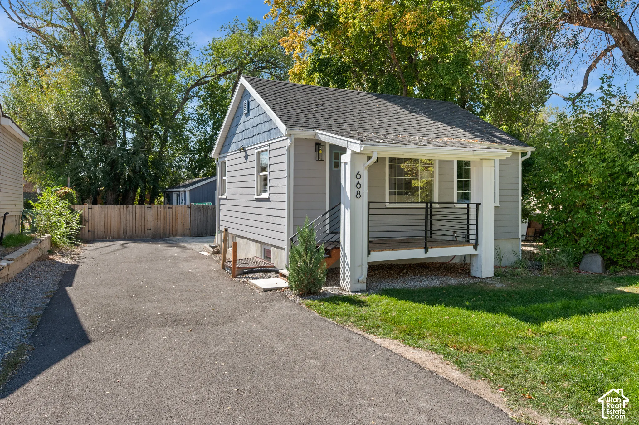 Gray siding exterior with blue shingle accent, covered porch with black railings, asphalt driveway, and a wood privacy fence