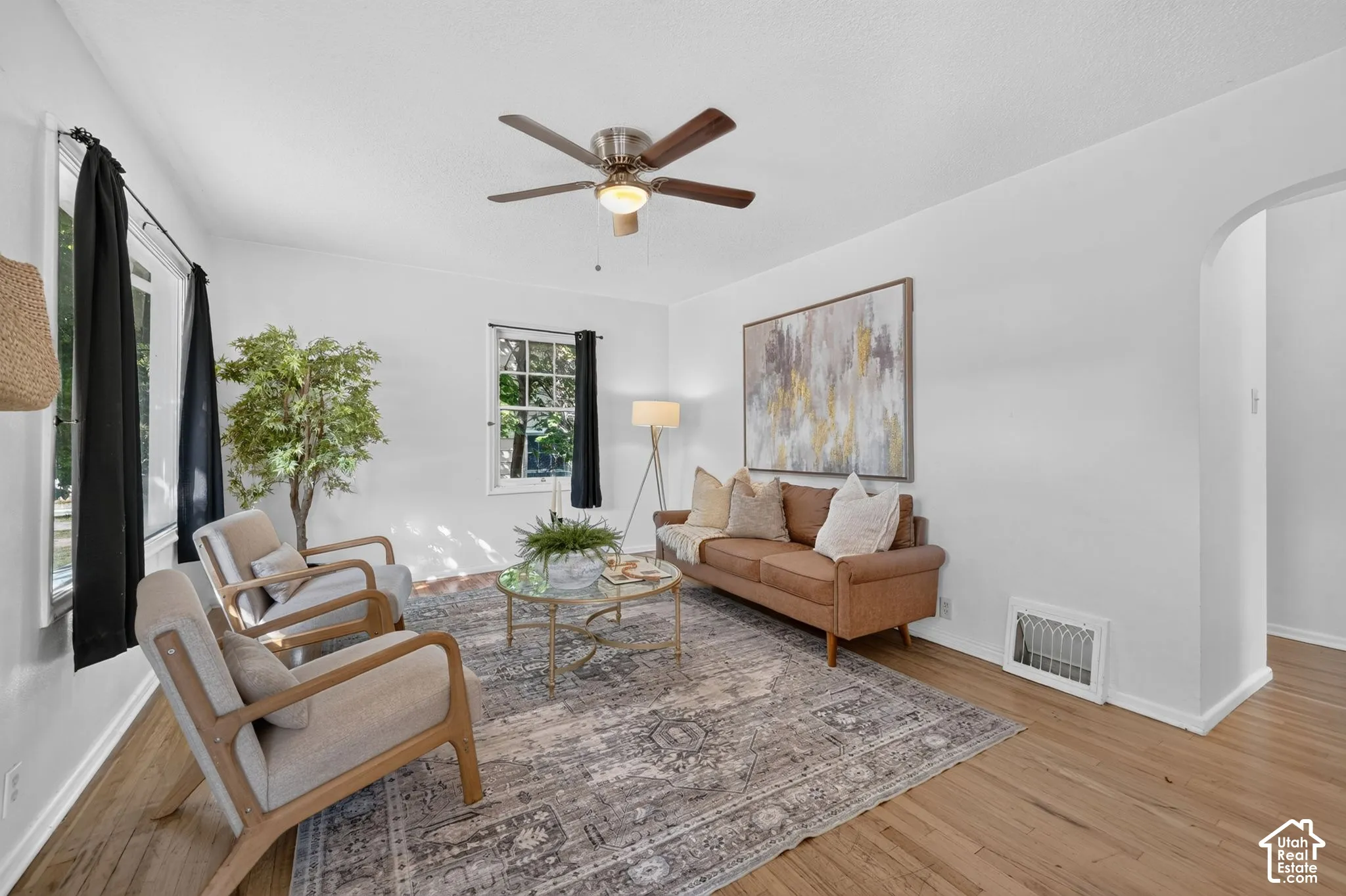 Hardwood flooring, white walls, multiple windows, ceiling fan, and an arched doorway