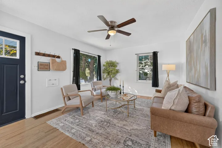 Hardwood flooring throughout, a dark paneled entry door with glass pane, bright white walls, a ceiling fan with light, and multiple windows with dark drapery rods