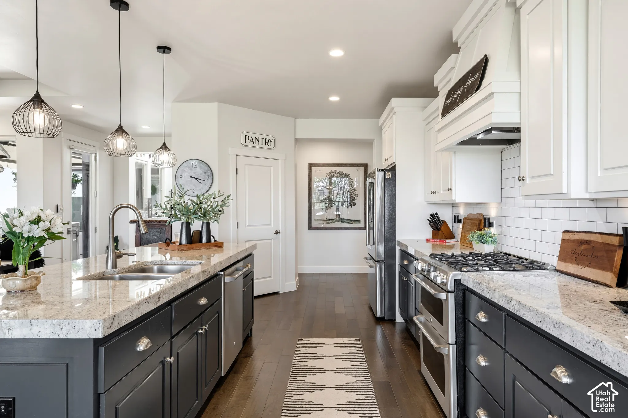 Galley-style kitchen featuring dual islands with light stone countertops, white cabinetry, stainless steel appliances including a gas range, pendant lighting, subway tile backsplash, and dark wood-finish flooring throughout the open layout
