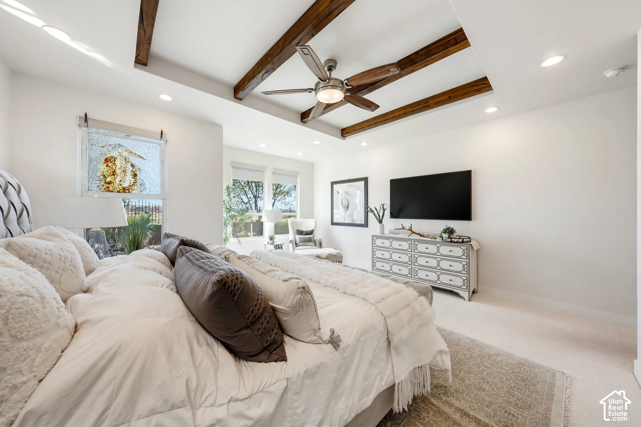 Spacious primary bedroom featuring exposed wood-beam ceiling detail, ceiling fan, wall-mounted flat-screen television, light wood-finish flooring, and an abundance of natural light from multiple windows