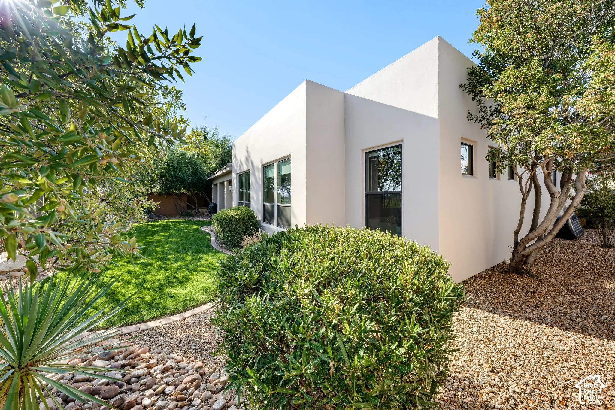 Contemporary stucco exterior with multiple windows, a manicured lawn, gravel landscaping, and mature trees