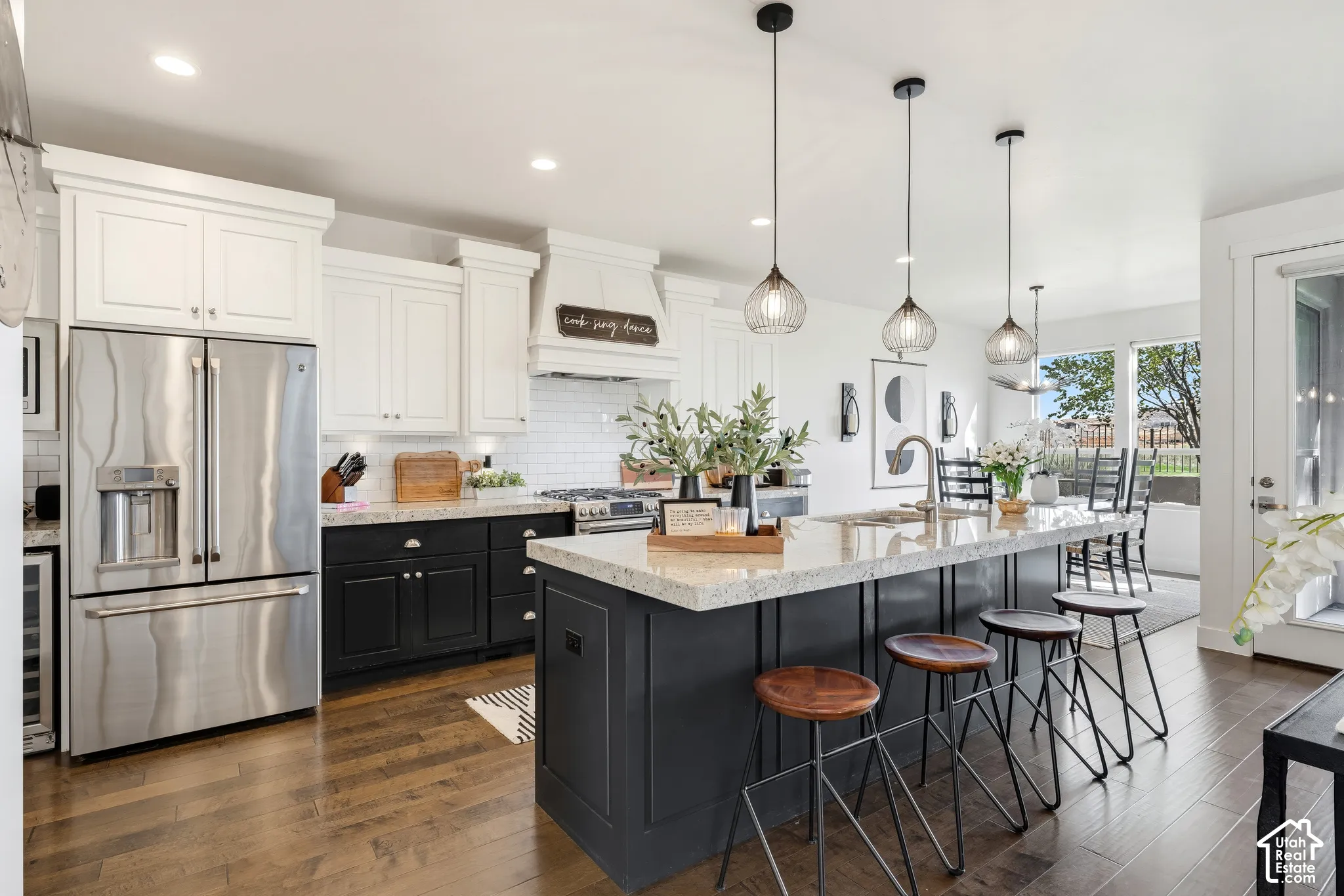 Gourmet kitchen featuring white cabinetry with brushed hardware, a black-finished island with light wood-finish countertops, stainless steel French-door refrigerator, pendant lighting with glass shades, and wood-finish flooring throughout