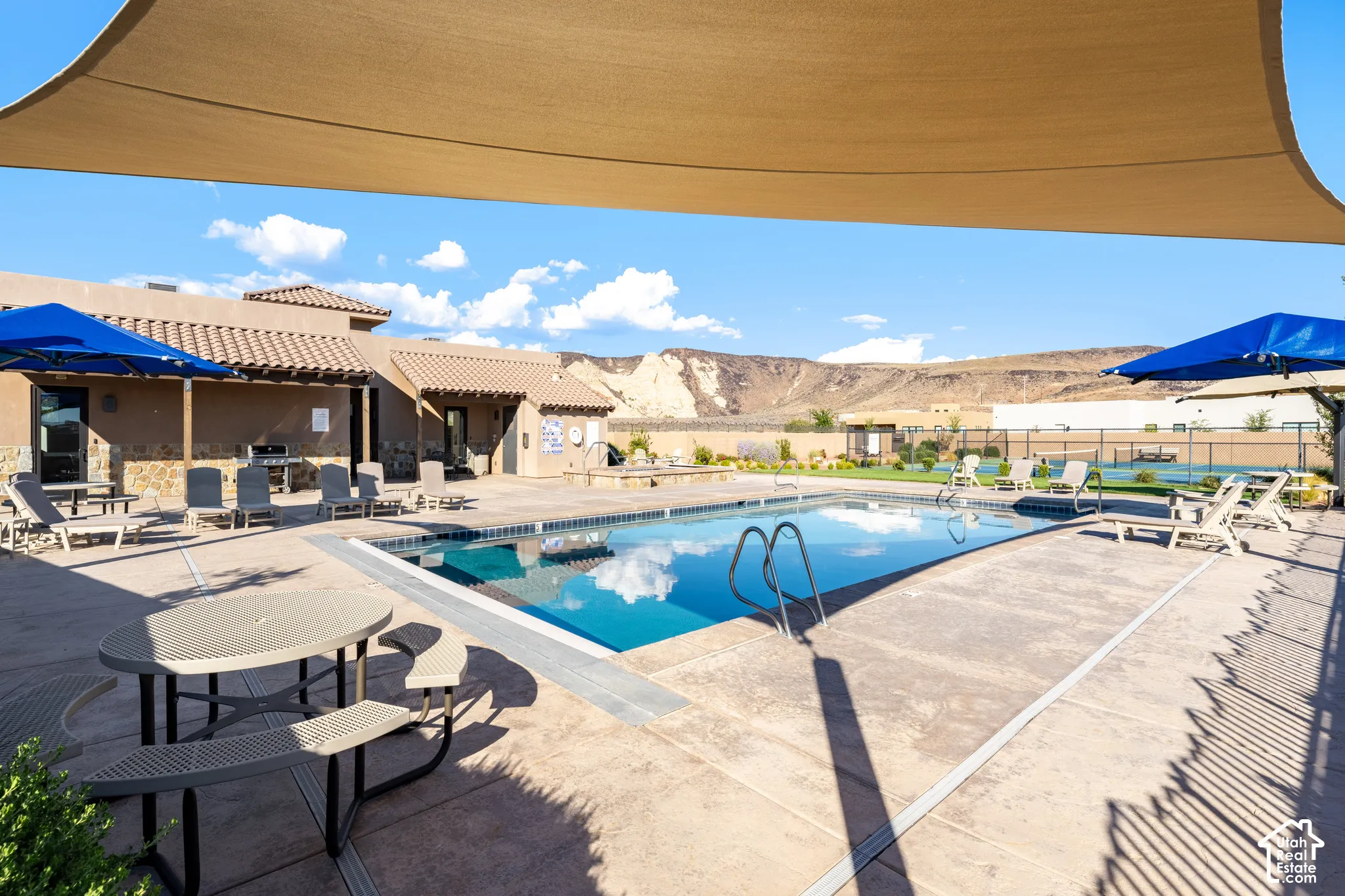 Resort-style pool area featuring crystal-blue water, concrete decking with integrated lounging spaces, multiple shade structures, and panoramic desert mountain backdrop