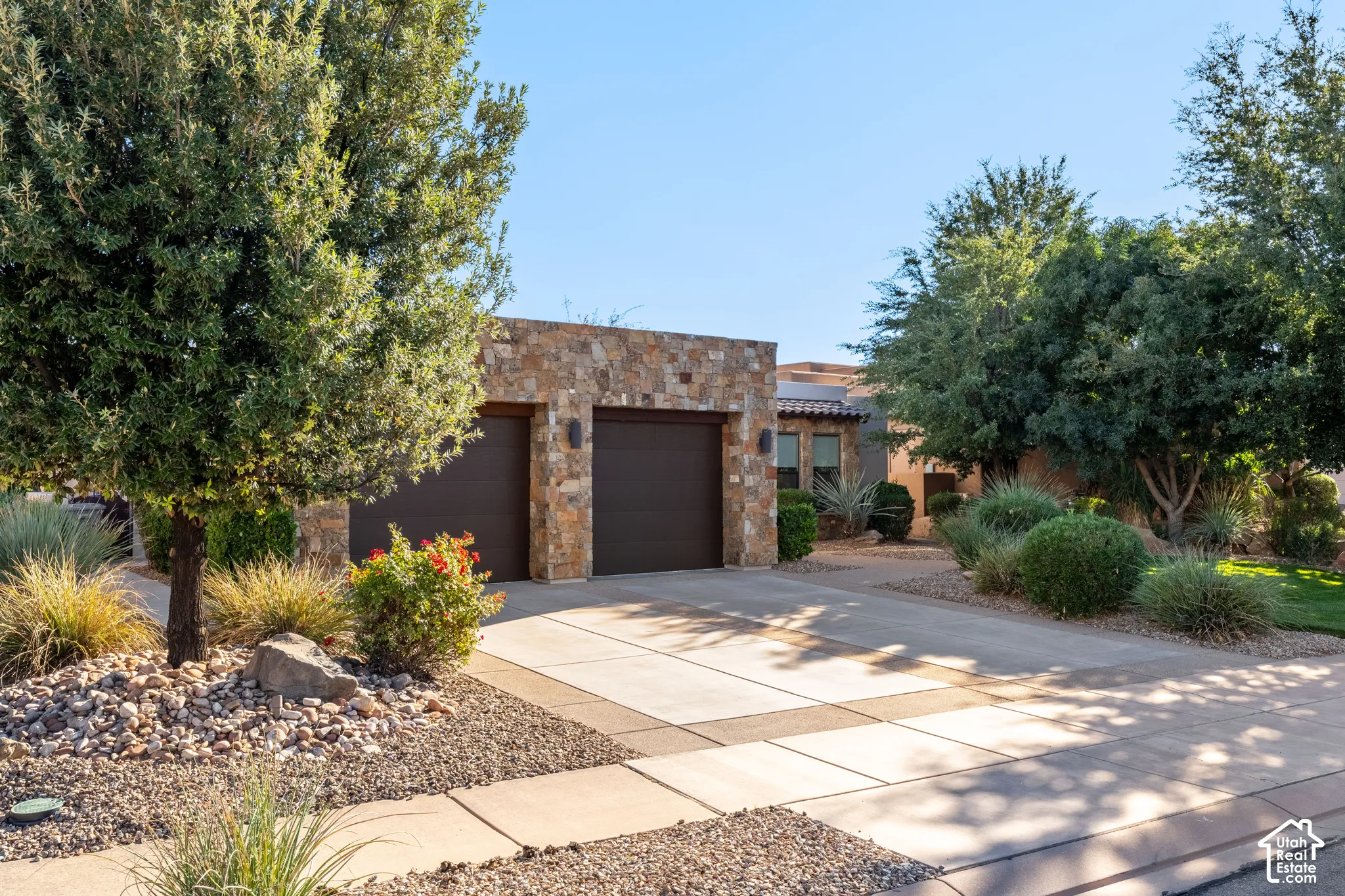 Stone veneer facade with integrated garage doors, concrete driveway, mature trees, and low-maintenance landscaping