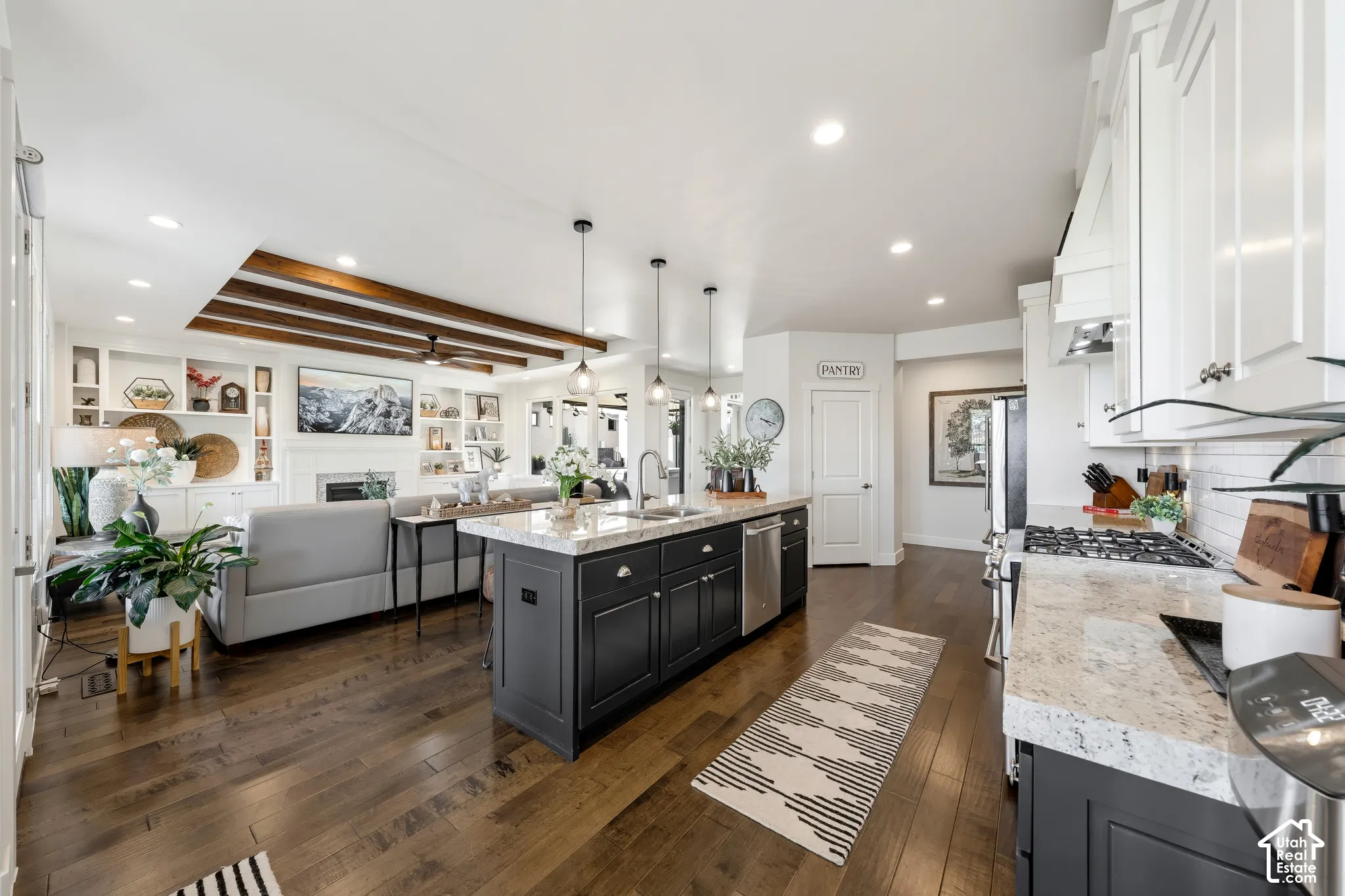 Open-concept kitchen featuring a large center island with black cabinetry, white perimeter cabinetry with marble countertops, stainless steel appliances, wood-finish flooring throughout, and recessed and pendant lighting with an architectural wood-beam ac