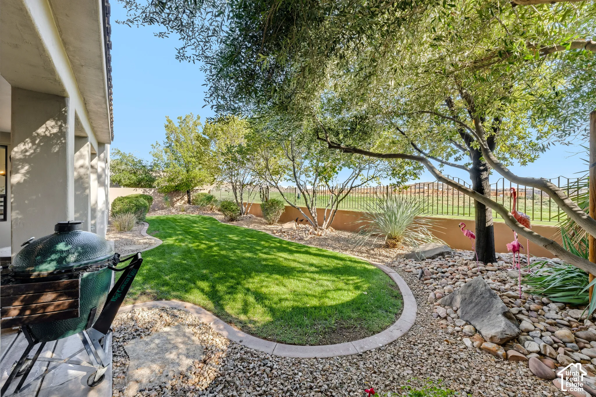 Expansive backyard oasis featuring a manicured lawn with curved hardscaping borders, mature shade trees, decorative rock landscaping, and metal fencing