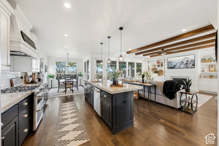 Open-concept kitchen and living area featuring a large island with dark cabinetry and wood-finish countertop, stainless steel appliances, white subway tile backsplash, exposed wood beam ceiling detail, hardwood flooring throughout, and abundant natural li