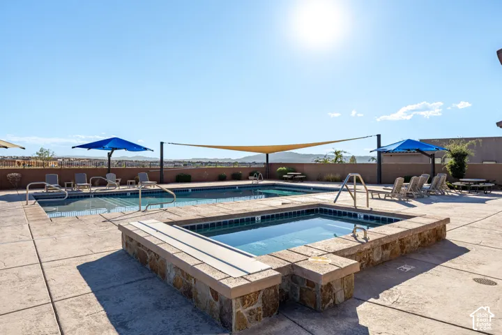 Resort-style outdoor living space featuring two swimming pools with stone decking, dual shade structures with blue umbrellas, and expansive hardscape patio