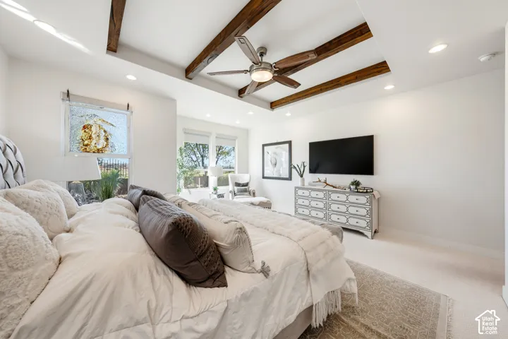 Spacious primary bedroom featuring exposed wood-beam ceiling detail, ceiling fan, wall-mounted flat-screen television, light wood-finish flooring, and an abundance of natural light from multiple windows