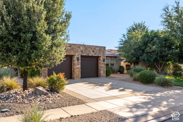 Stone veneer facade with integrated garage doors, concrete driveway, mature trees, and low-maintenance landscaping