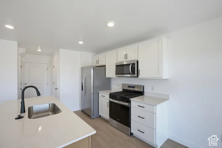 White cabinetry with black hardware, stainless steel appliances, wood-finish flooring, recessed lighting, and a kitchen island with an undermount sink