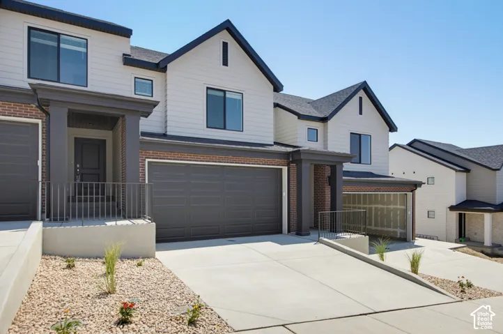 Modern multi-level townhome featuring a two-car garage, brick facade, covered porch entry, and dark-framed windows