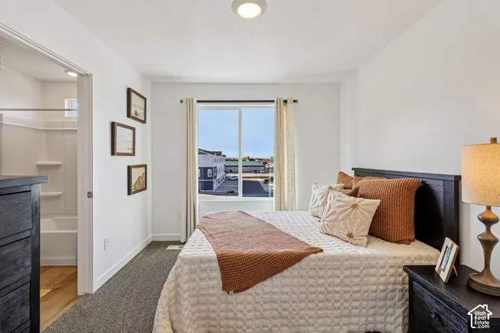 Bright primary bedroom featuring expansive windows with panoramic city outlooks, carpeted flooring, dark wood furnishings with built-in nightstands, warm ambient lighting via table lamp, framed wall art, and an ensuite bathroom accessible via adjacent doo