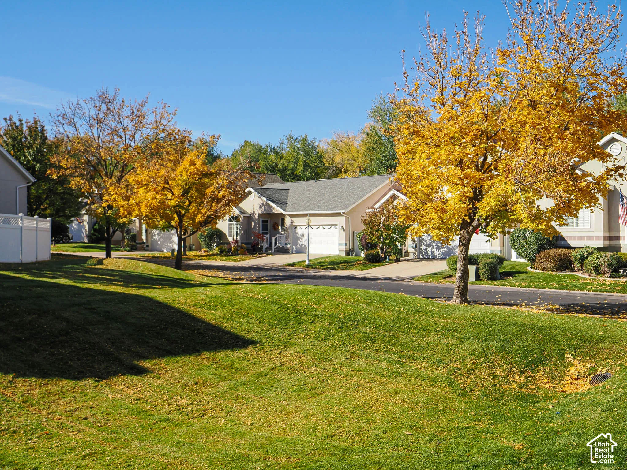 Manicured lawn, mature deciduous trees, asphalt driveway, two-car garage, and a privacy fence