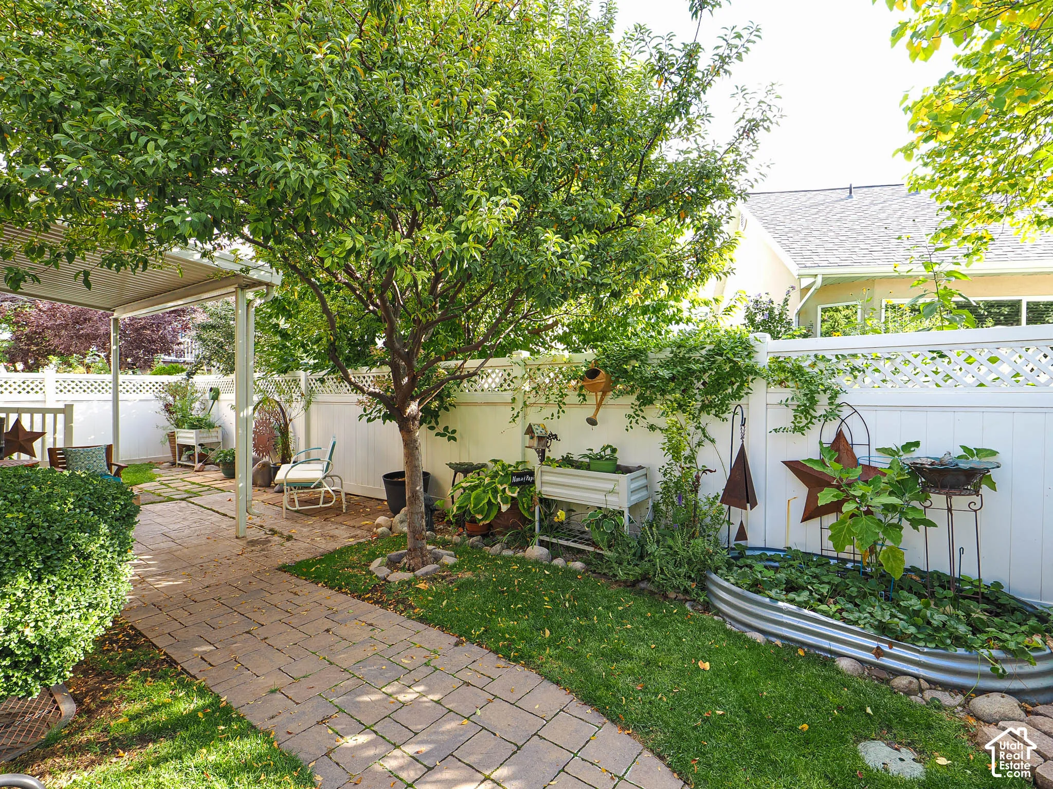 Covered patio with a paver walkway, manicured lawn, established landscaping, and white lattice fencing