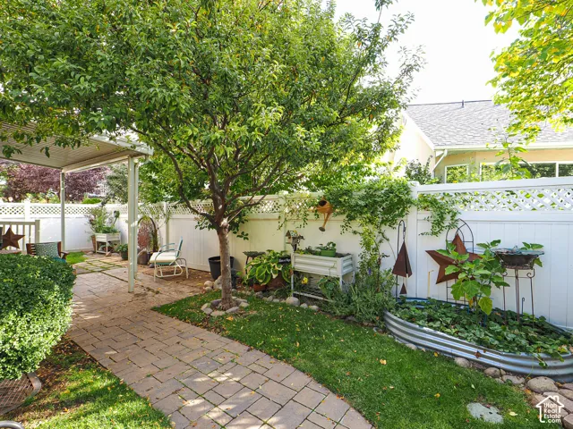 Covered patio with a paver walkway, manicured lawn, established landscaping, and white lattice fencing