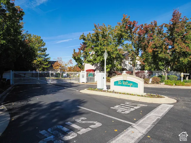 Gated community entrance featuring a stone sign with "The Village" lettering, mature trees, and a paved access road