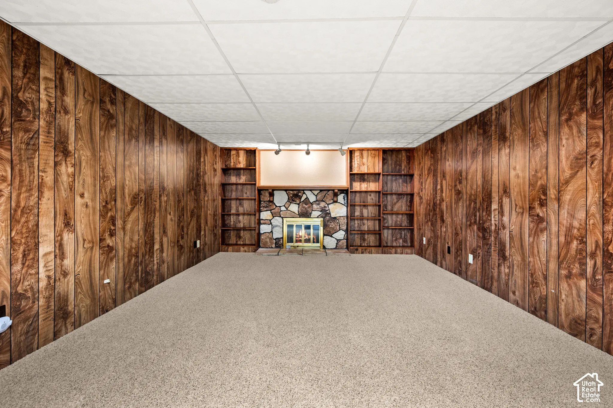Carpeted room featuring wood paneling, a stone fireplace, built-in shelving, and a drop ceiling with recessed lighting