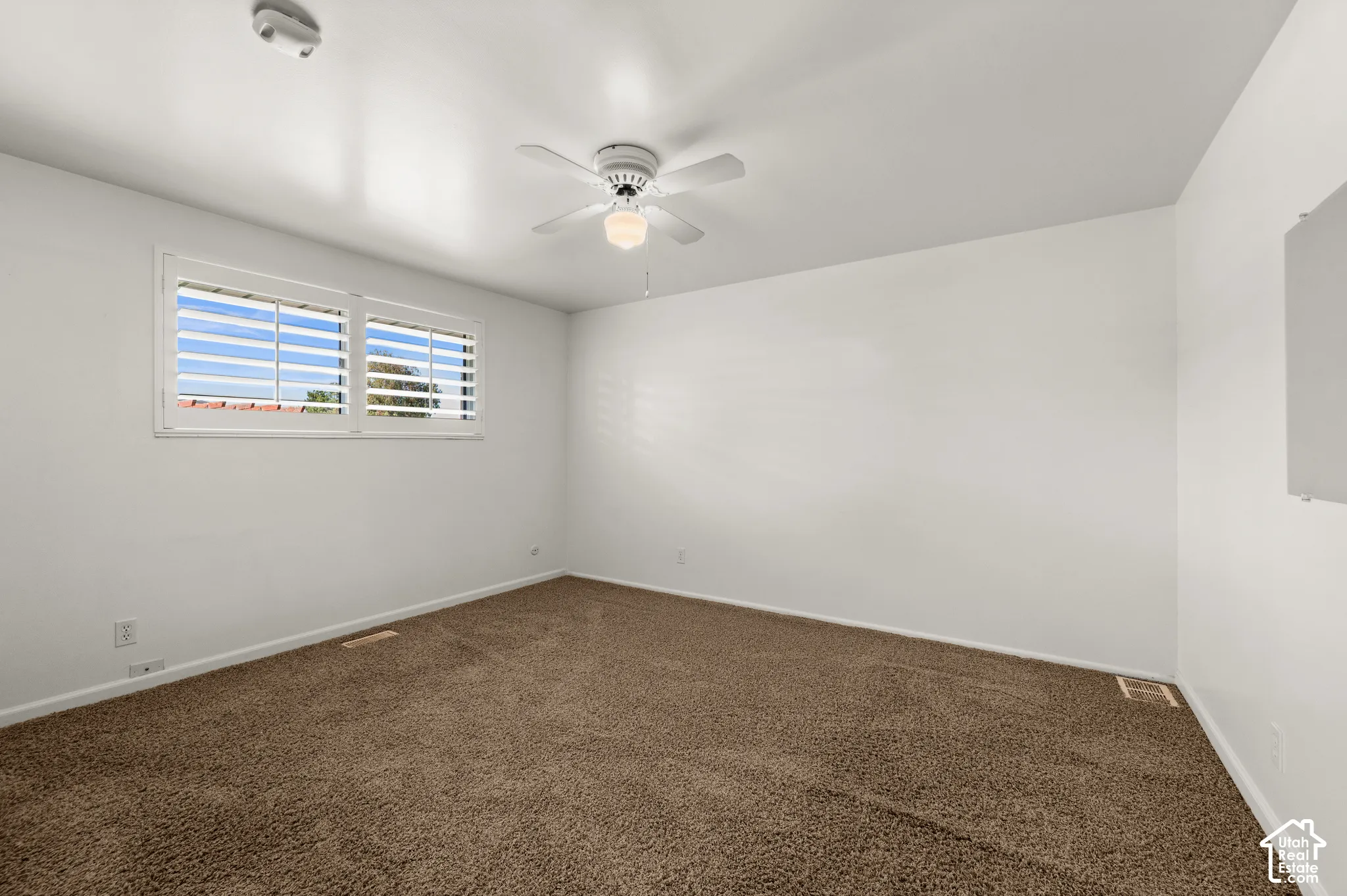 Room featuring soft brown carpeting, white walls, a white ceiling fan with integrated lighting, and a window with white louvered shutters
