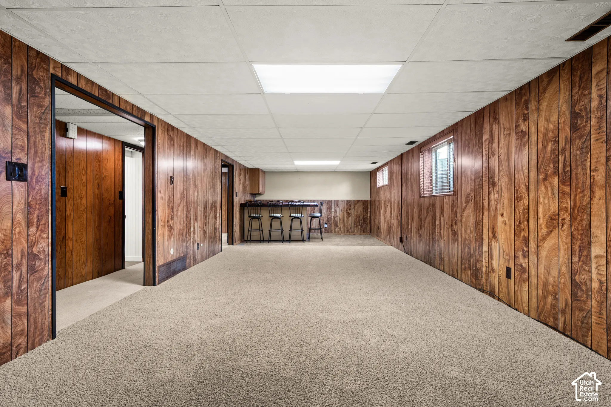 Finished lower level featuring wood-look paneling, drop ceiling with integrated lighting, carpeted flooring, and a built-in bar area with a countertop