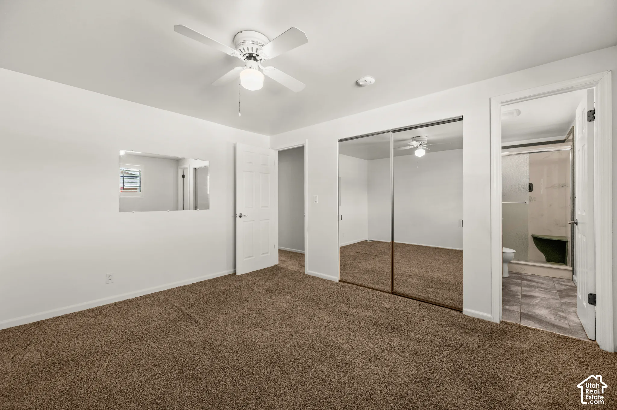 Brown carpet flooring, white walls, white ceiling fan with integrated lighting, mirrored sliding closet doors, and a paneled white door