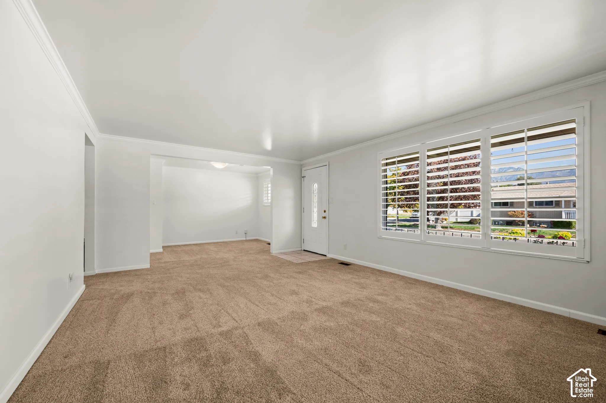 Spacious living area with neutral carpeting, white crown molding, and large windows featuring plantation shutters