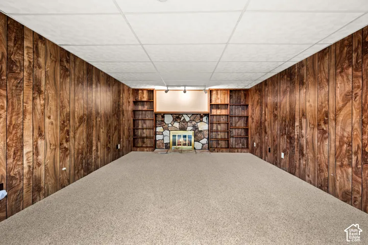 Carpeted room featuring wood paneling, a stone fireplace, built-in shelving, and a drop ceiling with recessed lighting