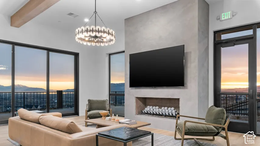 Expansive glass doors, floor-to-ceiling windows, an integrated fireplace, a modern chandelier, and exposed ceiling beams