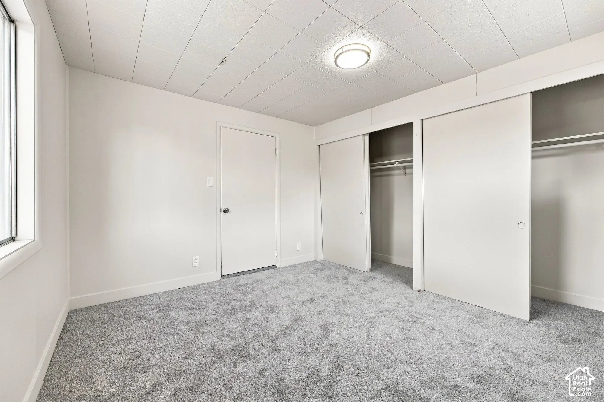 Bright room featuring light gray carpet flooring, a window, a flush-mount ceiling light fixture, and two sliding closet doors with built-in shelving