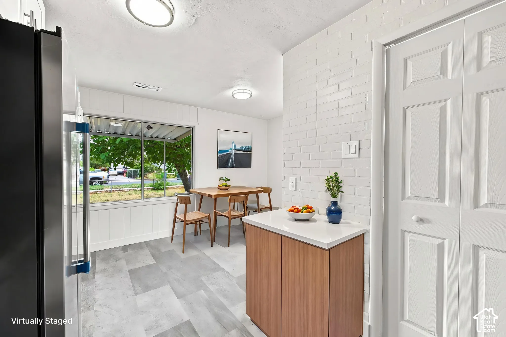 White painted brick accent wall, wood-finish cabinetry with white countertop, large window, and light-toned tile flooring