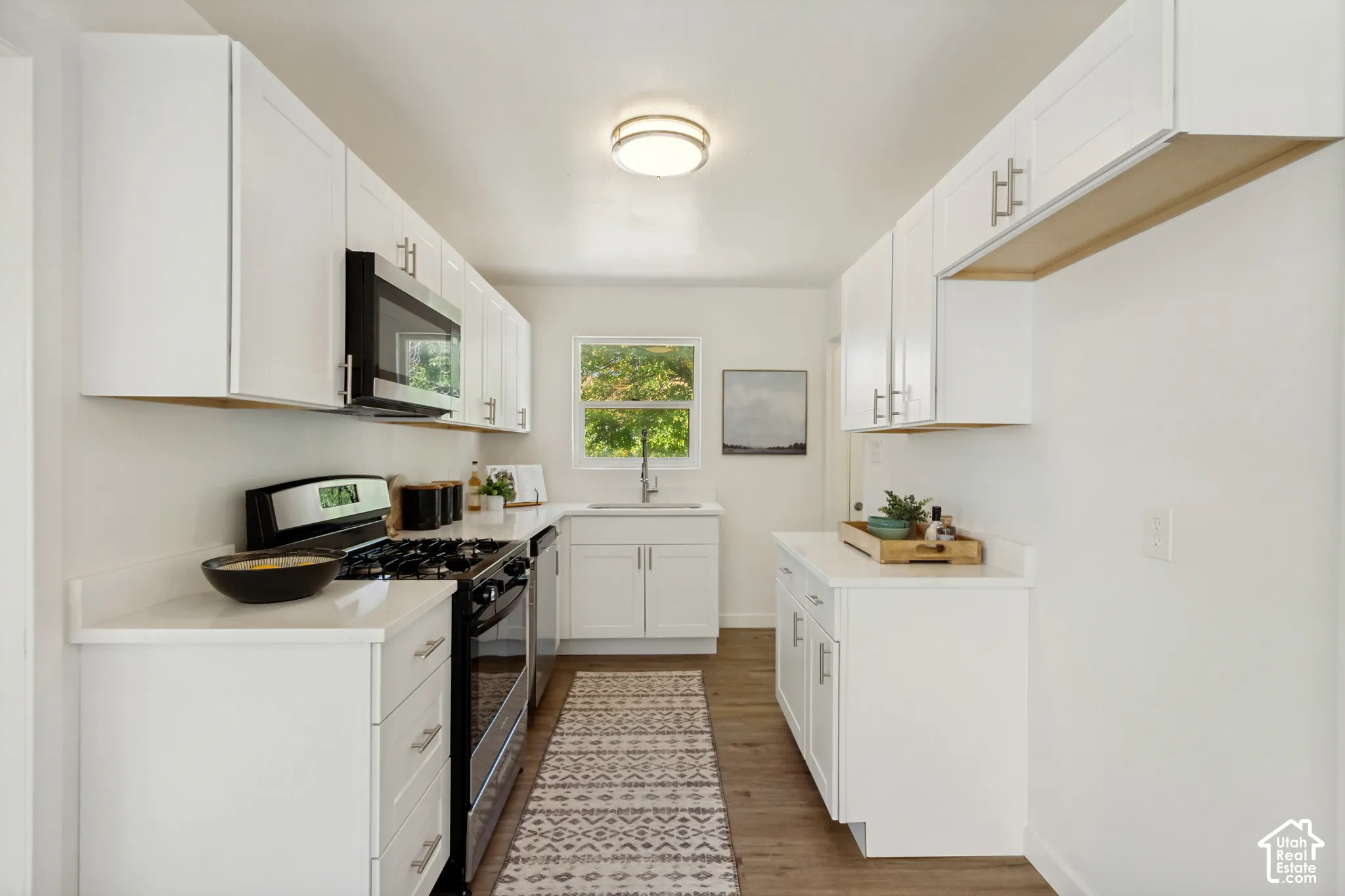 White shaker cabinetry, light-toned countertops, wood-finish flooring, stainless steel appliances, and a window over the sink