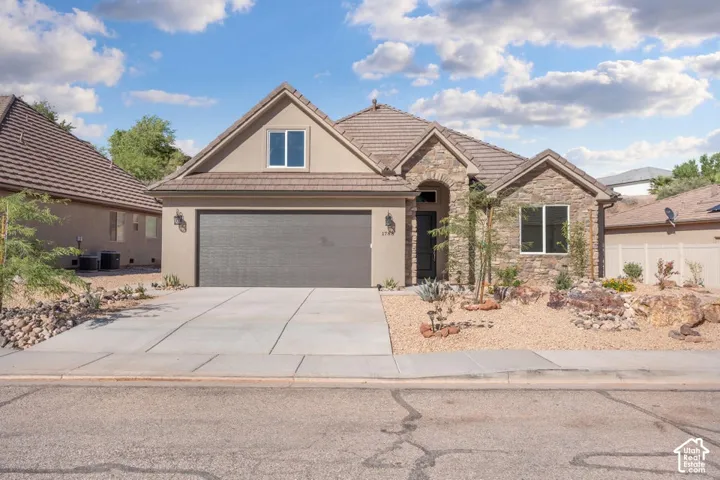 Stucco and stone facade with a tiled roof, featuring a two-car garage, mature desert landscaping, and a concrete driveway