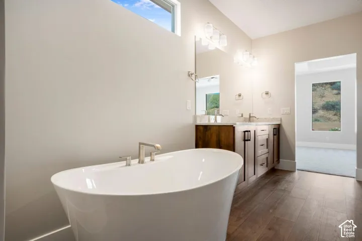 Freestanding soaking tub with brushed nickel fixtures, wood-finish flooring, dual vanity with light-toned countertop, and modern lighting fixtures