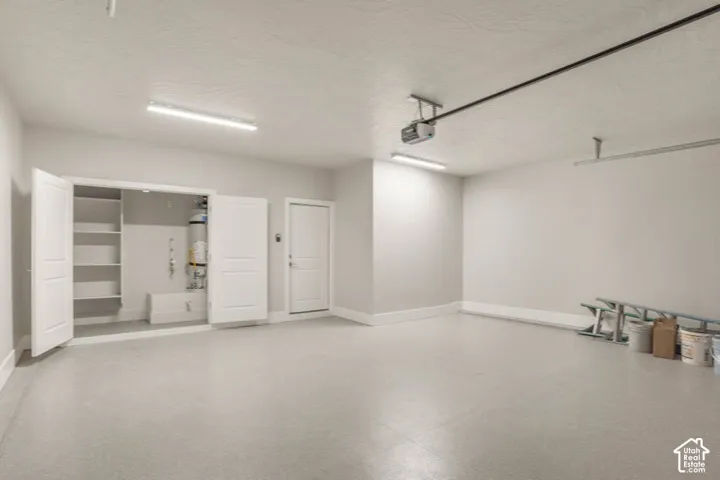 Spacious garage with light grey flooring, built-in shelving, two overhead fluorescent light fixtures, a ceiling-mounted garage door opener, and a white interior door