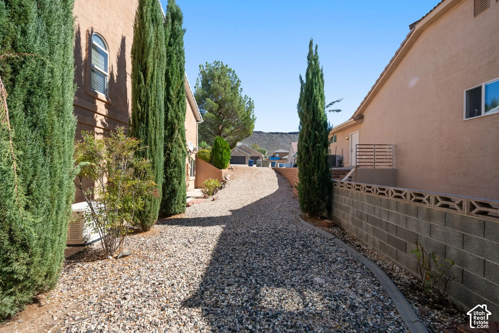 Gravel pathway bordered by established evergreen trees and a decorative block retaining wall