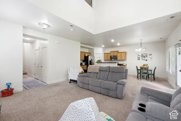 Open-concept living space featuring high ceilings, light-colored carpeting, a kitchen with wood-finish cabinetry, a breakfast bar, and a dining area with a chandelier
