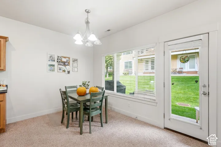 Carpeted dining area featuring a multi-light chandelier, a large rectangular window with blinds, and a glass-paneled door