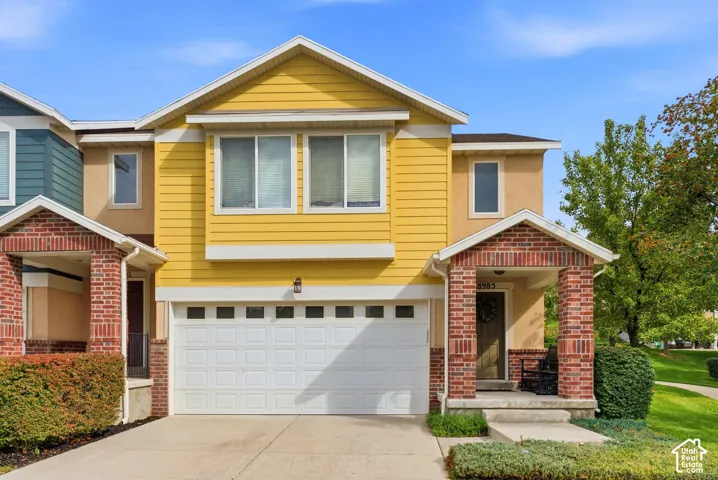 Yellow siding exterior with white trim, a two-car garage, a brick-clad covered porch, and a concrete driveway