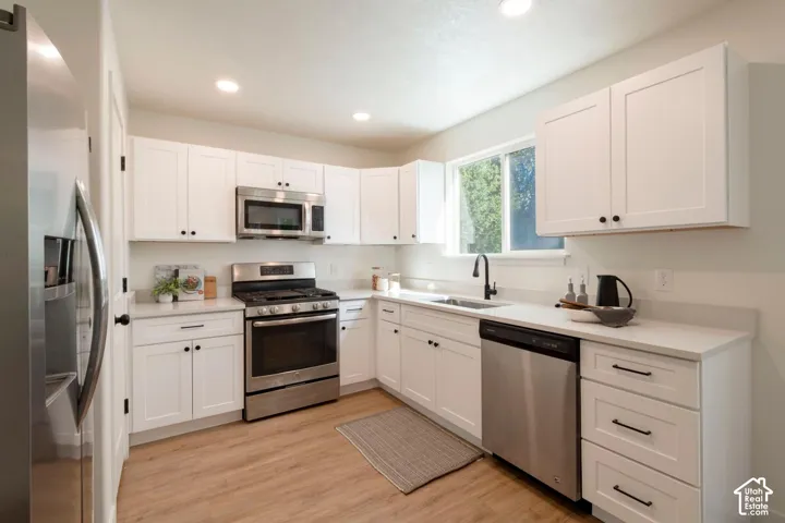 Modern kitchen featuring white shaker-style cabinetry, stainless steel appliances, light-toned countertops, wood-finish flooring, and a black gooseneck faucet
