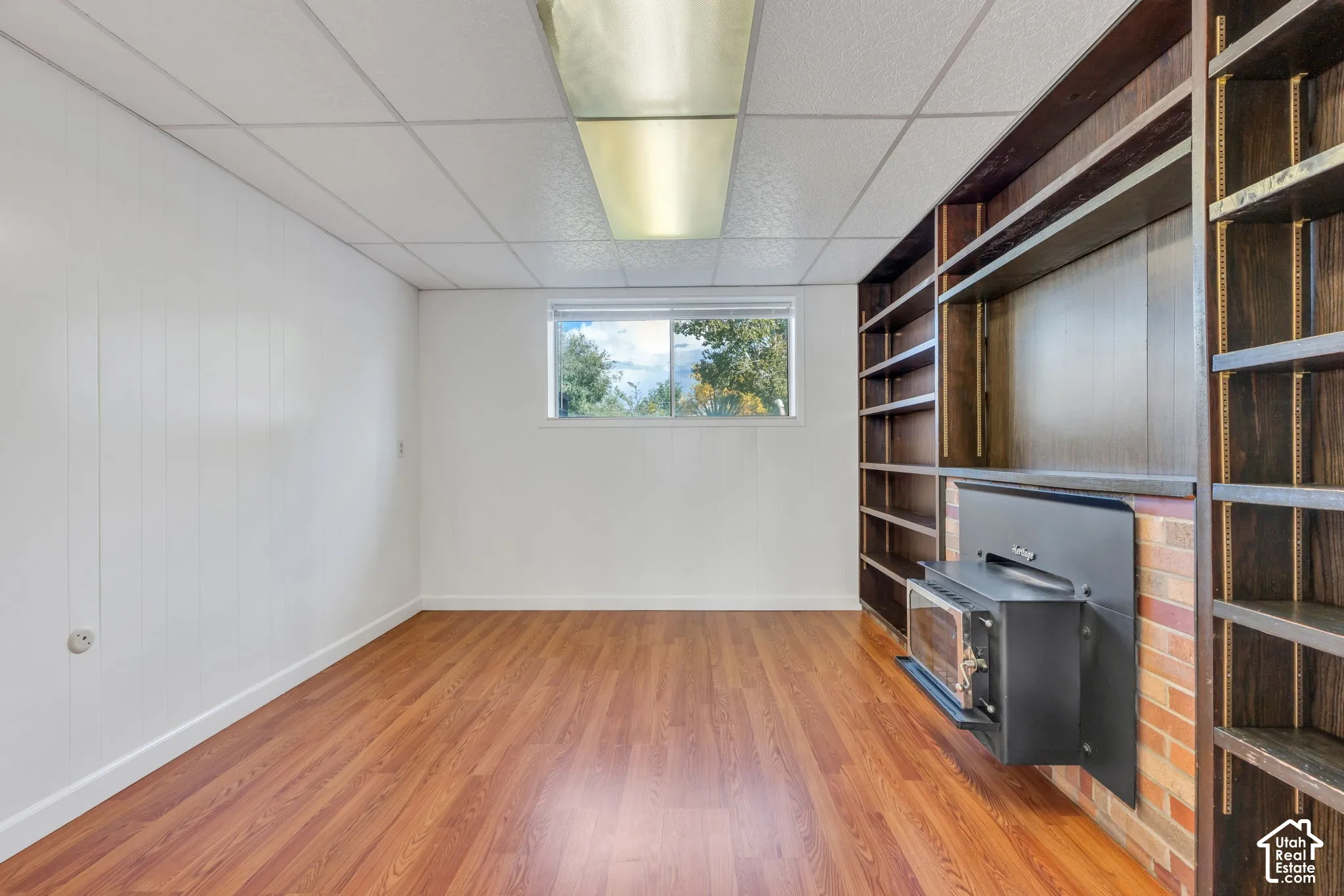 Wood-finish flooring, built-in shelving, fireplace insert with brick surround, and a drop ceiling with fluorescent lighting