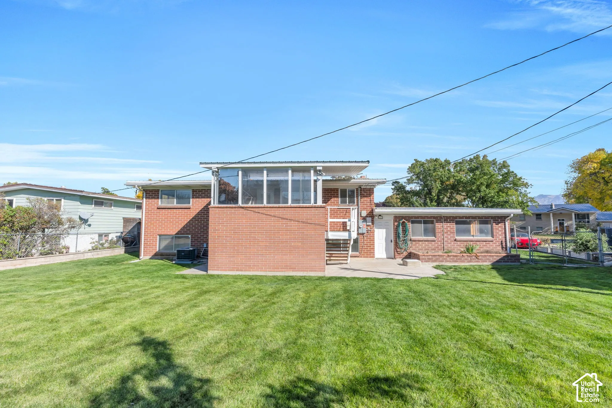 Expansive green lawn, brick exterior, upper-level enclosed sunroom, concrete patio, and exterior stairs